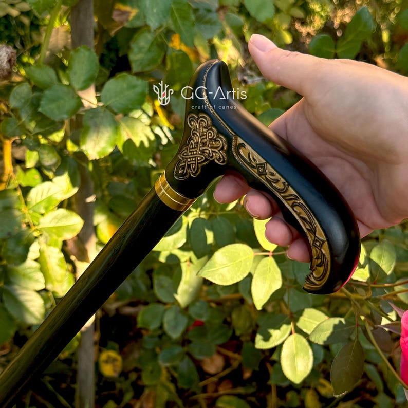 Outdoor photo of the Celtic cane placed among sunlit green leaves. The handle’s gold engraving and polished finish stand out against the bright foliage. The black shaft adds a clean vertical line, presenting the cane as a stylish, handcrafted accessory suited for outdoor or everyday settings.