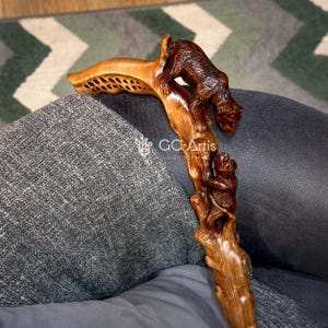 May include: A wooden walking cane with intricate carvings of a bear and a monkey. The handle has a unique, open-work design. The cane is made of light and dark brown wood, with the text "GC-Artis" visible.