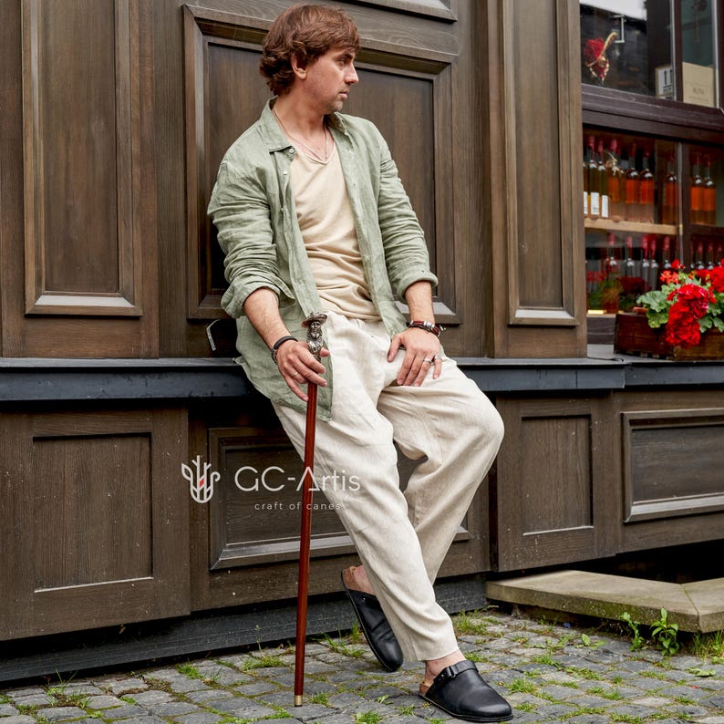 May include: A man wearing a green linen shirt and beige pants is sitting on a wooden ledge. He is holding a wooden walking stick with a decorative metal top. The stick is about 40 inches long.