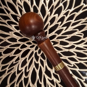 May include: A wooden walking stick with a rounded knob handle and a gold band. The stick is brown and has a smooth finish. The text "GC-Artis craft of canes" is visible on the stick.