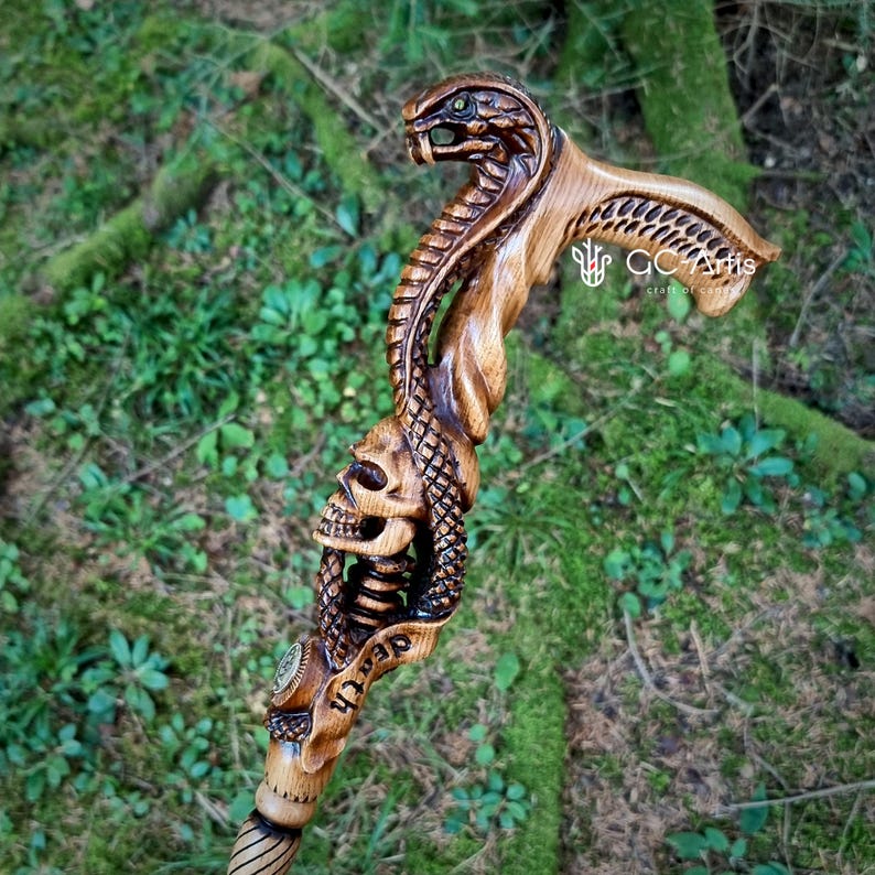 Hand carved wooden walking cane with intertwined snake and skull motif photographed outdoors on green moss. Detailed carving highlights coiled serpent body wrapping around the shaft and skull design. GC-Artis cane blends gothic fantasy art with functional support and collectible craftsmanship.