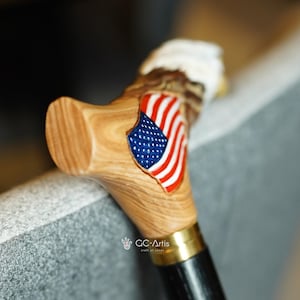 May include: A wooden walking cane handle featuring a carved eagle head with a white top and a polished light brown wood grain finish. An American flag emblem with red, white, and blue colors is inlaid on the side. A gold metal collar connects the handle.