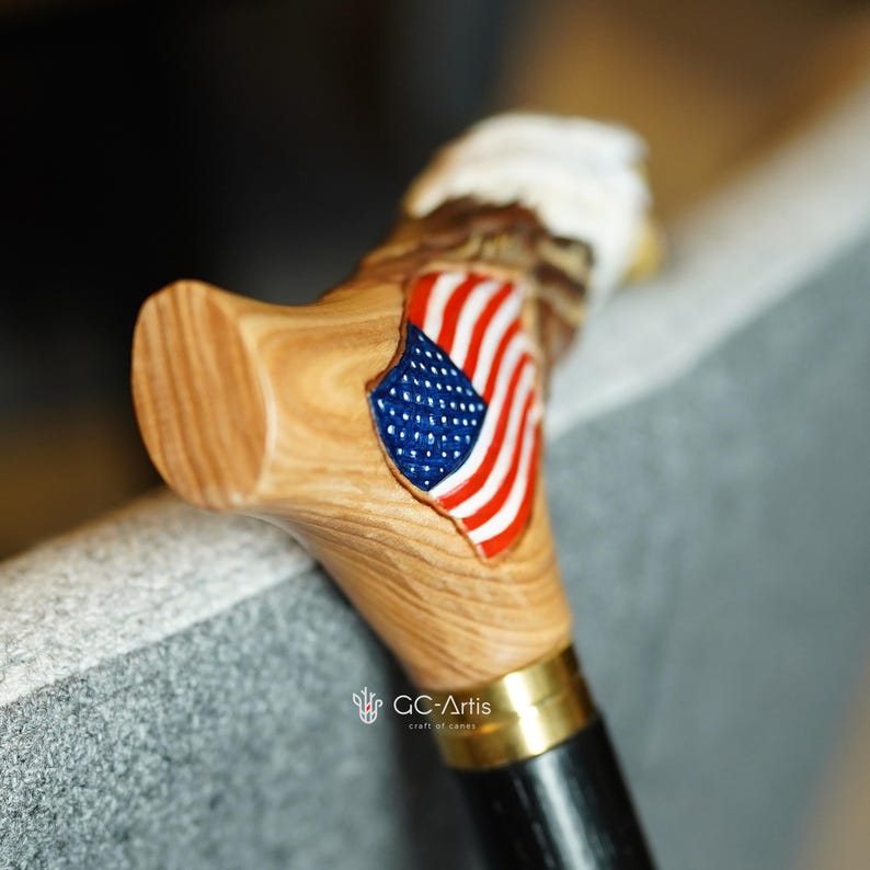 May include: A wooden walking cane handle featuring a carved eagle head with a white top and a polished light brown wood grain finish. An American flag emblem with red, white, and blue colors is inlaid on the side. A gold metal collar connects the handle.
