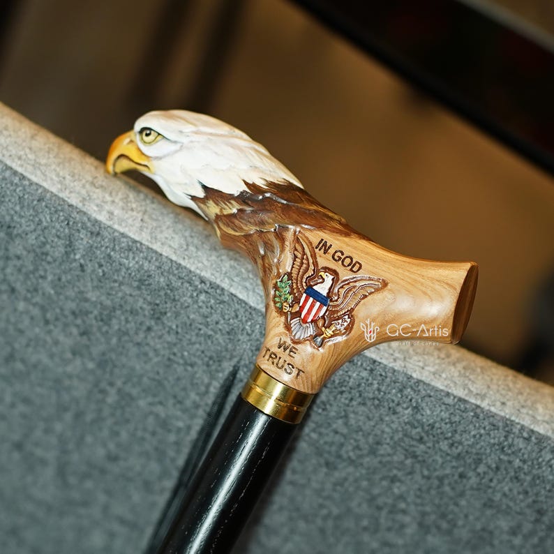 May include: A walking cane featuring a carved wooden handle shaped like a bald eagle head with a natural wood finish. The handle displays a carved eagle emblem with the text IN GOD WE TRUST and a gold-colored metal collar above a black shaft.