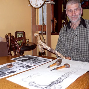 May include: A man sits at a table with a wooden walking stick, carving tools, and sketches of the stick. The stick is intricately carved with a detailed design.