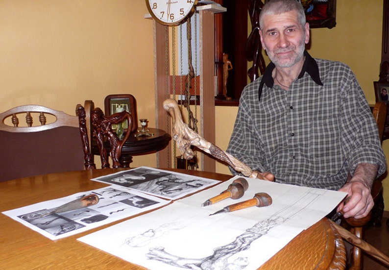 May include: A man sits at a table with a wooden walking stick, carving tools, and sketches of the stick. The stick is intricately carved with a detailed design.