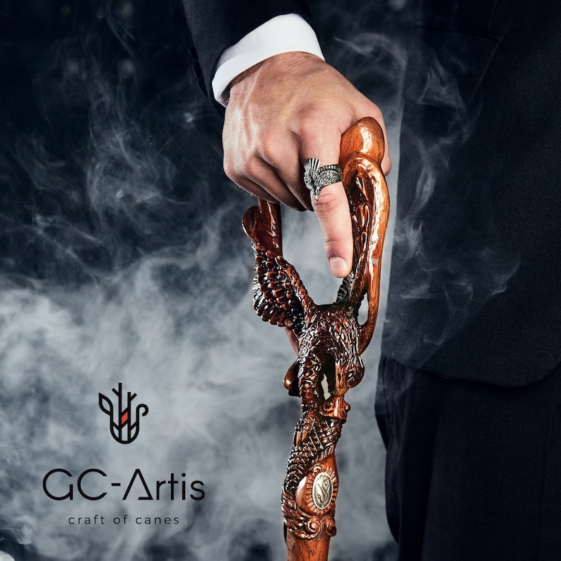 May include: A wooden walking stick with a carved eagle design. The stick is held by a hand wearing a silver ring with a black design. The background is a blurry image of smoke.