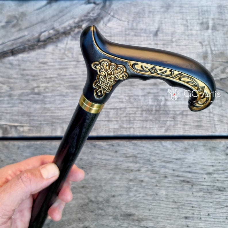 Outdoor photo of the Celtic cane held near a blooming pink rose. The engraved handle with gold accents contrasts with the bright flower, emphasizing craftsmanship and decorative detail. The black shaft frames the scene, showing how the cane looks in the hand during real outdoor use.