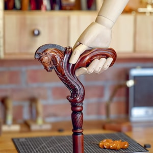 May include: A dark brown wooden walking cane featuring a detailed hand-carved horse head handle with a mane and bridle design. The shaft of the cane has a smooth, cylindrical finish with a decorative twisted pattern just below the horse head handle.