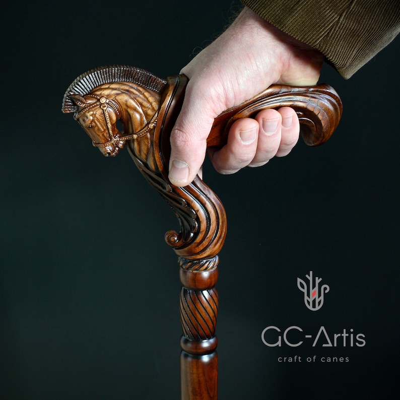 May include: A dark brown wooden walking cane featuring a detailed hand-carved horse head handle with a mane and bridle. The shaft has a spiral-carved section below the handle. The logo GC-Artis craft of canes is visible in the bottom right corner.