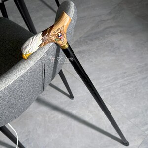 Full side view of the bald eagle cane leaning on a modern chair. The black shaft and golden brass collar create elegant contrast with the painted wooden handle. GC-Artis cane stands for balance between modern design and heritage symbolism.