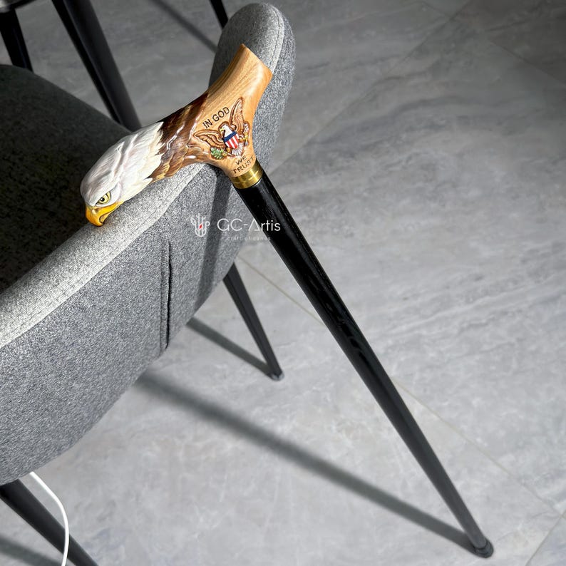 Full side view of the bald eagle cane leaning on a modern chair. The black shaft and golden brass collar create elegant contrast with the painted wooden handle. GC-Artis cane stands for balance between modern design and heritage symbolism.