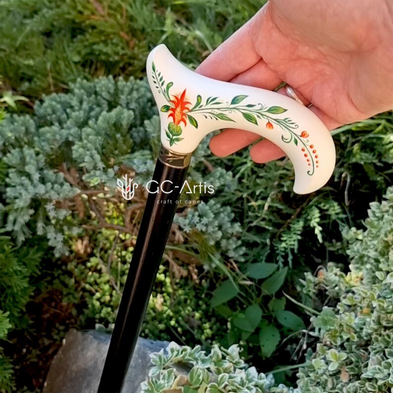 Puede incluir: Un bast&oacute;n blanco con un dise&ntilde;o floral pintado a mano con flores naranjas y verdes. El bast&oacute;n tiene un eje de madera negro y una punta de metal dorada. El texto "GC-Artis craft of canes" es visible en el eje.