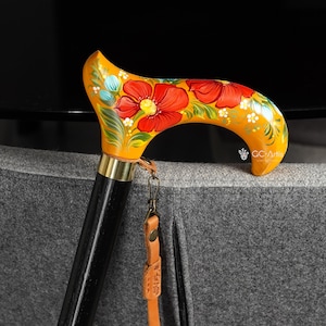 Può includere: Un bastone da passeggio in legno con un'impugnatura curva, dipinto di giallo e decorato con motivi floreali rossi. L'impugnatura ha un collare in ottone e un fusto nero. È attaccato un cinturino in pelle con chiusura in metallo.