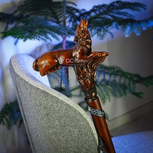 May include: A wooden walking stick with a carved wolf head handle. The wolf is howling and has a detailed, realistic design. The stick is brown and has a black strap around it. The text "GC-Artis craft of carve" is visible on the strap.