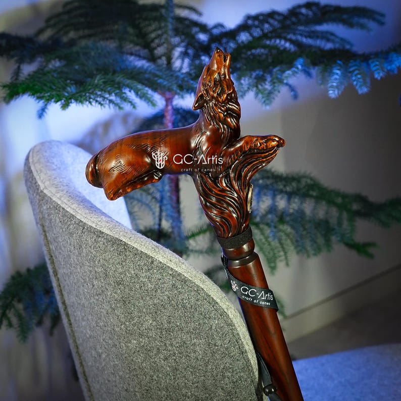 May include: A wooden walking stick with a carved wolf head handle. The wolf is howling and has a detailed, realistic design. The stick is brown and has a black strap around it. The text "GC-Artis craft of carve" is visible on the strap.