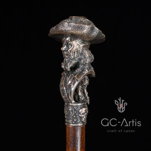 May include: A silver walking stick with a detailed bust of a pirate wearing a hat. The bust is attached to a wooden shaft with a skull and crossbones design.