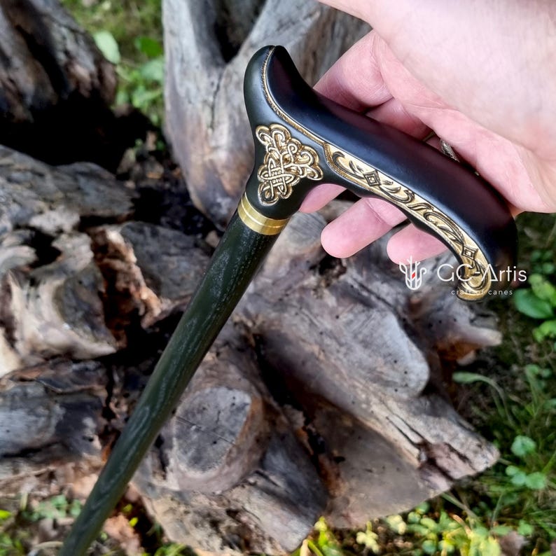 Outdoor photo of the Celtic cane resting on natural driftwood. The engraved handle stands out with gold accents and an elegant curved shape. The black shaft leans against the textured wood, creating a contrast between handcrafted precision and rugged natural surfaces. The image shows the cane in a nature-inspired setting, emphasizing its artistic detail.