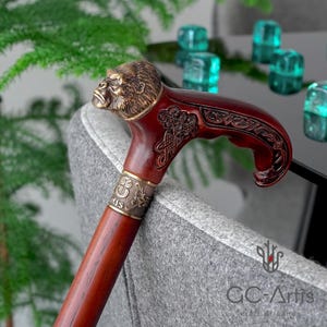 May include: A wooden walking stick with a carved gorilla head handle. The handle is made of brass and has intricate details. The stick is brown and has a brass band with the word "MIS" engraved on it.
