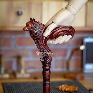 May include: A wooden walking stick with a carved wolf head handle. The handle is intricately detailed and features a flowing design. The stick is brown and has a twisted shaft.