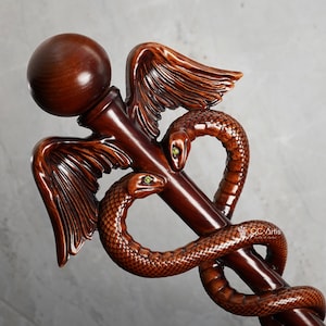 May include: A polished dark brown wooden caduceus staff featuring a central rod topped with a sphere, flanked by two carved wings and two intertwined snakes with green eyes. The text CC Artis is visible on the lower right side of the carving.