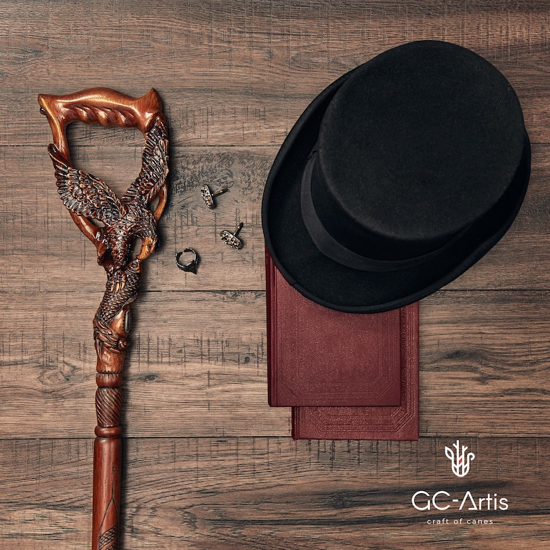 May include: A wooden walking stick with an eagle carving, a black top hat, two cufflinks, a ring, and two books on a wooden surface. The text "GC-Artis craft of canes" is visible in the bottom right corner.