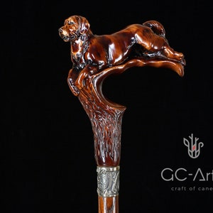 May include: A wooden walking stick with a detailed carving of a brown dog leaping from a tree branch. The stick has a silver band with an engraved design.