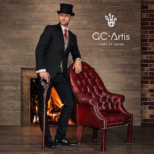 May include: A man in a black suit and top hat stands next to a red leather armchair. He is holding a wooden walking stick with a decorative top. The background is a brick wall with a fireplace and a sign that reads "GC-Artis craft of canes".