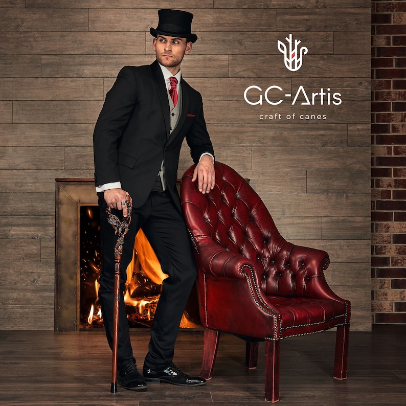 May include: A man in a black suit and top hat stands next to a red leather armchair. He is holding a wooden walking stick with a decorative top. The background is a brick wall with a fireplace and a sign that reads "GC-Artis craft of canes".