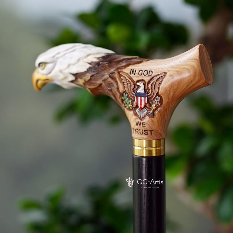 May include: A walking cane featuring a carved wooden handle shaped like a bald eagle head with a painted eagle emblem and the text IN GOD WE TRUST. The handle is attached to a black wooden shaft by a gold-colored metal collar, showcasing detailed craftsmanship.