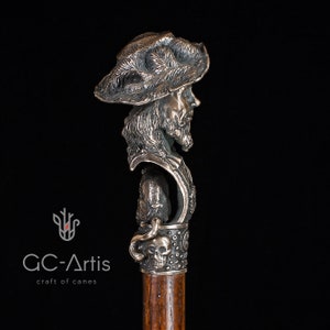 May include: A silver walking stick with a detailed sculpted head of a man wearing a hat. The stick has a skull and crossbones design on the handle.