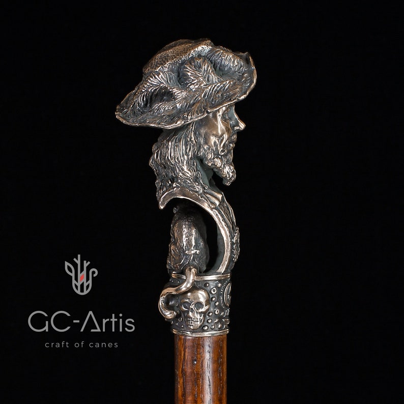 May include: A silver walking stick with a detailed sculpted head of a man wearing a hat. The stick has a skull and crossbones design on the handle.