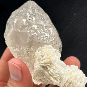 May include: A clear quartz crystal with a pointed tip, connected to a cluster of off-white, textured mineral formations. The quartz is translucent, and the mineral cluster has a rough, layered texture.