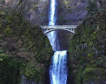 May include: A scenic view of a waterfall cascading down a rocky cliff. A white arched bridge spans the waterfall, surrounded by lush green vegetation. The water is a bright blue, contrasting with the dark rocks and greenery.