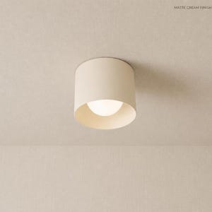 以下が含まれることがあります： マットクリーム仕上げの円筒形フラッシュマウント天井照明器具。この器具は頑丈な外装ハウジングと中央の円形開口部を備えており、そこから柔らかな光を放つ丸みを帯びた白い電球が見えています。