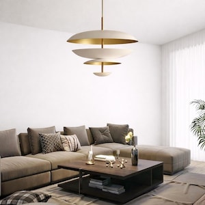 May include: A contemporary pendant light with a cream exterior and gold interior, suspended from a gold rod. The light fixture is above a living room with a brown sectional sofa and a dark wood coffee table. The light has a tiered design.