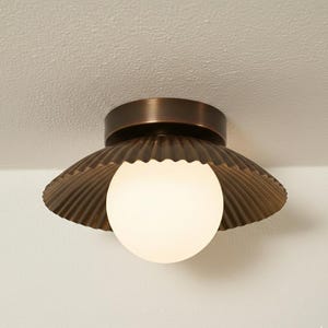 May include: A bronze ceiling light with a scalloped shade and a white globe. The light is mounted on a white ceiling. The design is reminiscent of a seashell. The fixture is a decorative lighting element.