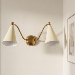 May include: A brass wall light with two cream-coloured, cone-shaped shades. The fixture has curved arms extending from a circular base. It is mounted on a light-coloured wall, with a framed artwork visible in the background.