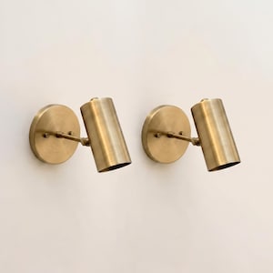 Può includere: Due applique da parete color ottone. Ogni applique presenta una base circolare, un braccio sottile e un apparecchio di illuminazione cilindrico. Gli apparecchi sono leggermente angolati verso il basso. Le applique sono montate su una parete bianca.