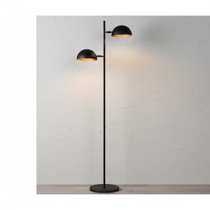 May include: A black metal floor lamp with two dome-shaped light fixtures. The interior of the lampshades is gold. The lamp has a tall, slender pole and a round base. The background is a light-coloured wall.