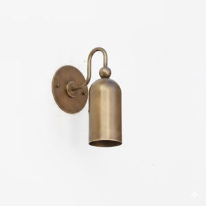 May include: A brass wall sconce featuring a bell-shaped shade and a curved arm. It has a circular backplate and a warm, metallic finish. The design is simple and elegant, suitable for various interior styles. The sconce is approximately 20 cm in height.