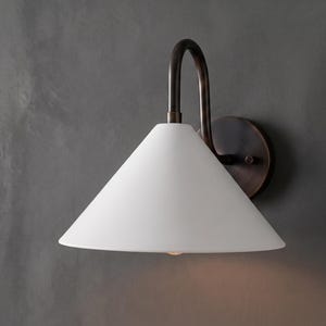May include: A wall sconce featuring a conical white glass shade attached to a dark bronze metal arm. The fixture includes a circular wall mounting plate and a curved neck that supports the downward-facing shade, emitting a soft warm light.