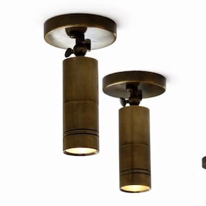 May include: Two bronze-coloured, cylindrical ceiling spotlights with adjustable heads. Each light has a circular base and a small, round light at the bottom. The lights are suspended against a white background.