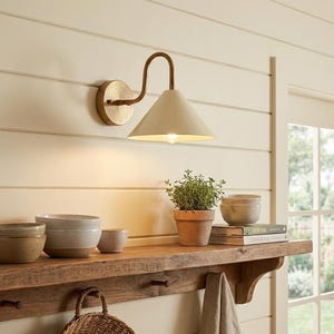 May include: A wall-mounted lamp with a curved brass arm and a conical off-white shade. The fixture is attached to a circular brass base and emits a warm light downward, illuminating a wooden shelf holding ceramic bowls and a potted plant.