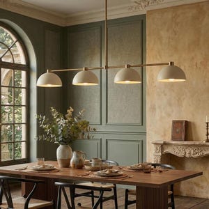 May include: A contemporary four-light pendant lamp with a brass finish and off-white dome shades hangs above a wooden dining table. The room features a green wall with paneling and a window with a view of greenery.