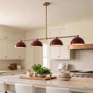 May include: A linear chandelier featuring a long, thin brass rod suspended from the ceiling, holding four dome-shaped pendant lights in a deep burgundy finish. Each dome has a white interior, providing downward-facing illumination.