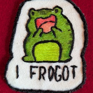 Puede incluir: Parche bordado con una rana verde cubriéndose la cara. La rana está sobre un fondo blanco con un borde negro. El texto "I FROGOT" está debajo en letras negras. Ideal para ropa o accesorios.