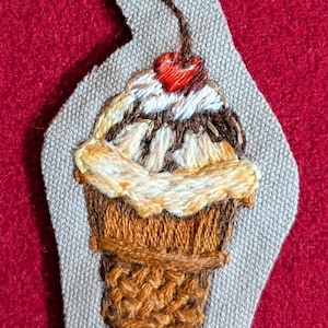 Puede incluir: Parche bordado que representa un helado sundae detallado. El helado tiene un cono marrón, helado de vainilla y chocolate, sirope de chocolate y una cereza roja encima. El parche está sobre un fondo gris con un fondo de fieltro rojo.