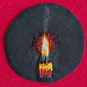 Può includere: Toppa ricamata con un disegno a candela. La fiamma è nei toni del rosso, arancione, giallo e bianco, con dettagli blu. La candela è su uno sfondo circolare nero, con un sbuffo di fumo bianco. Oggetto decorativo.