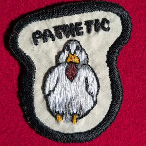 May include: Embroidered patch with the word "PATWETIC" above a cartoon bird figure. The bird is white with a red necktie and yellow feet. The patch has a black border and is set against a red background.